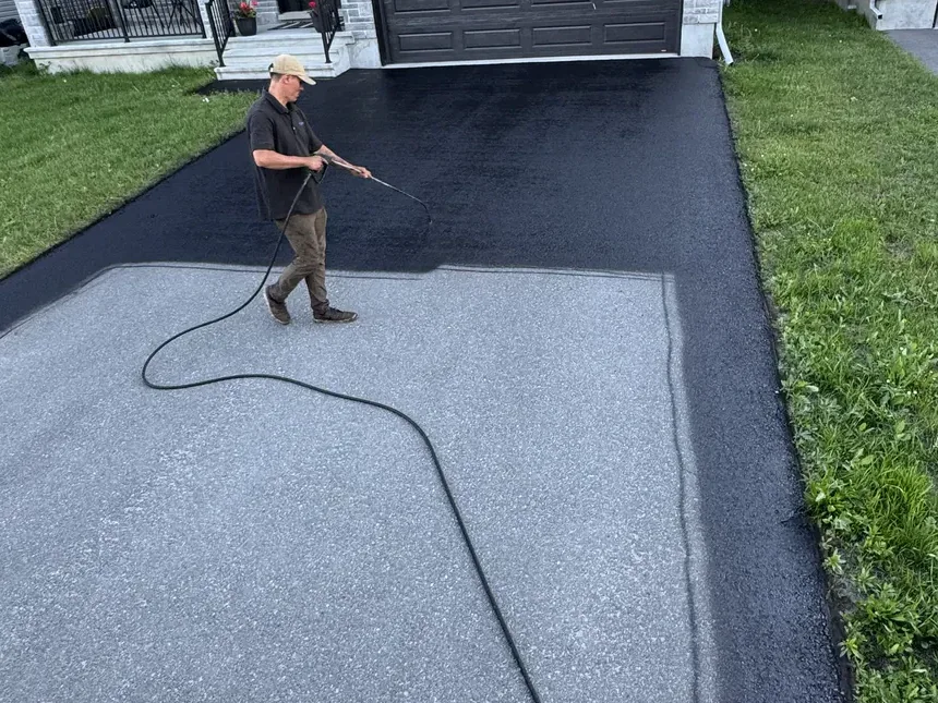 Worker spraying dark sealer along the edge of a residential driveway.